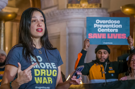 A DPA staff member leading a rally in Albany, NY. Her shirt reads "No More Drug War." A sign reads "Overdose Prevention Centers Save Lives".