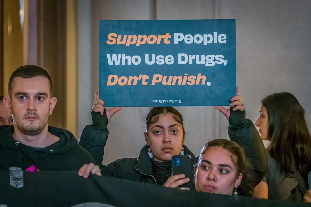 A woman holds a sign reading "Support People Who Use Drugs, Don't Punish."