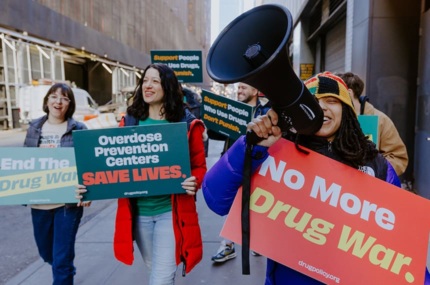 A diverse group of people protest on a NYC sidewalk with a megaphone. Their signs read "No More Drug War," "Overdose Prevention Centers Save Lives", "End the Drug War", and "Support People Who Use Drugs, Don't Punish."