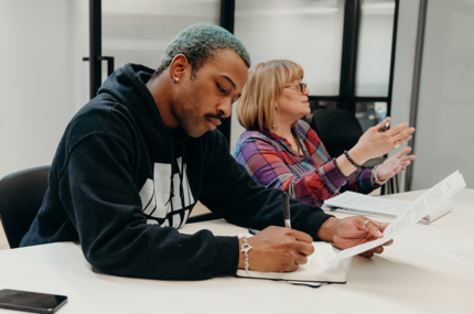 Two people at a meeting. A young man writes in a notebook, consulting pages of notes, while a woman speaks to someone else in the background.