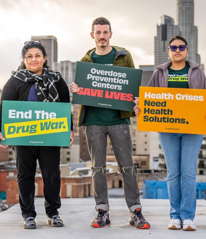 Three people stand in front of the LA cityscape holding signs reading "End the Drug War", "Overdose Prevention Centers Save Lives", and "Health Crises Need Health Solutions".