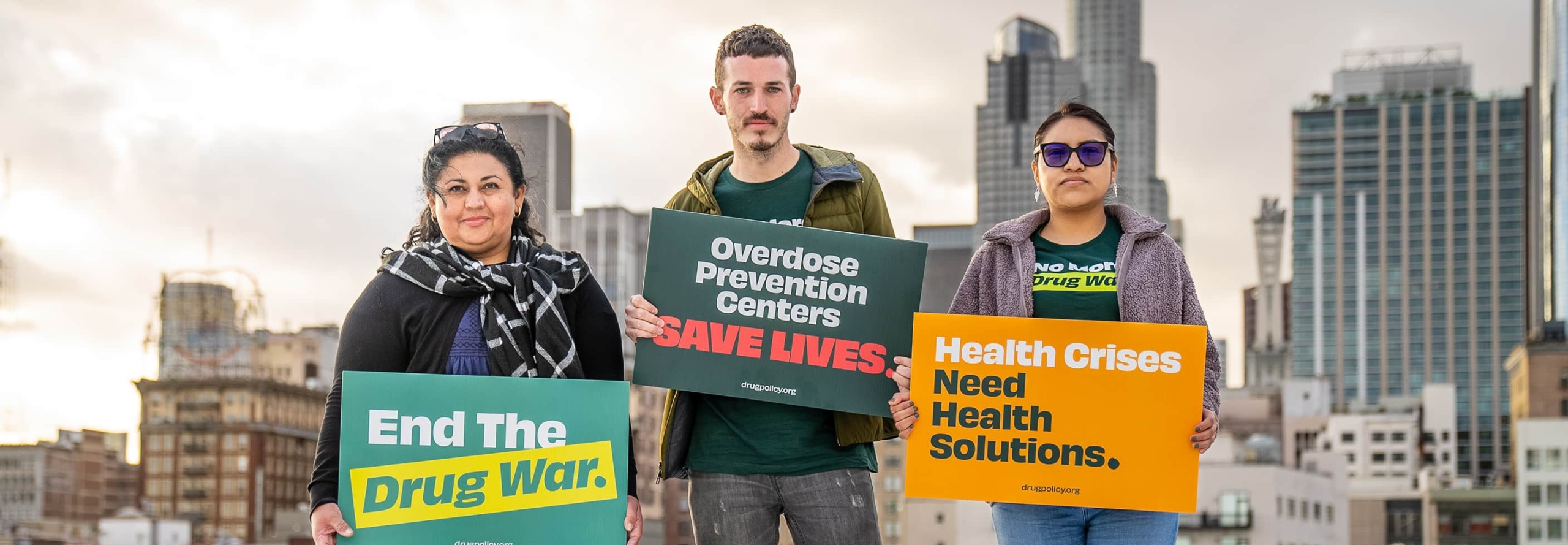 Three people stand in front of the LA cityscape holding signs reading "End the Drug War", "Overdose Prevention Centers Save Lives", and "Health Crises Need Health Solutions".