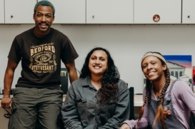 Three diverse young people sit in an office, smiling at the camera.