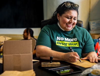 Latina woman wearing a shirt reading "No More Drug War" writes a message on a paper bag at a volunteer outreach center.
