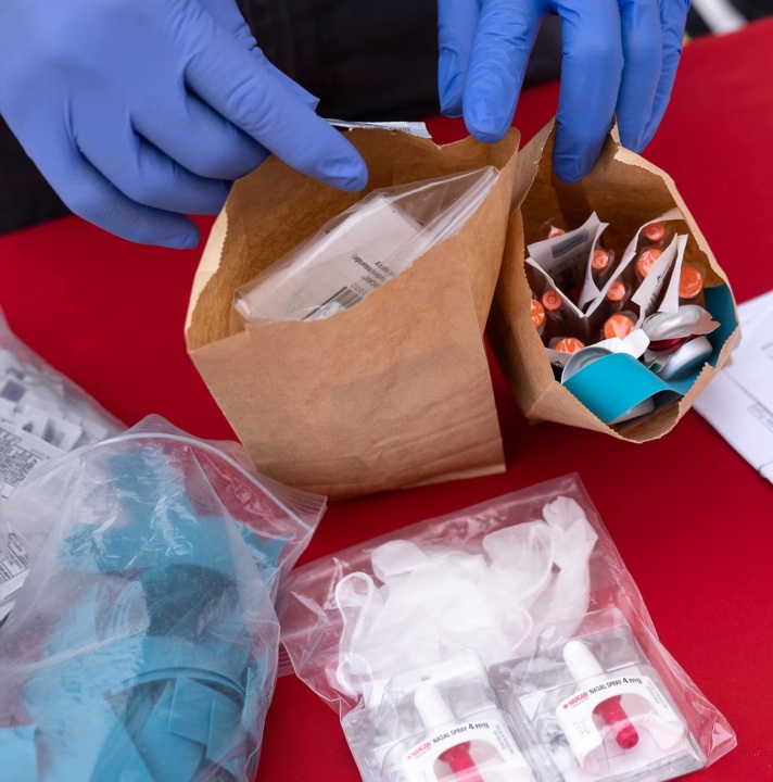 Hands in medical gloves show paper bags full of harm reduction supplies on a table, including clean supplies and naloxone.