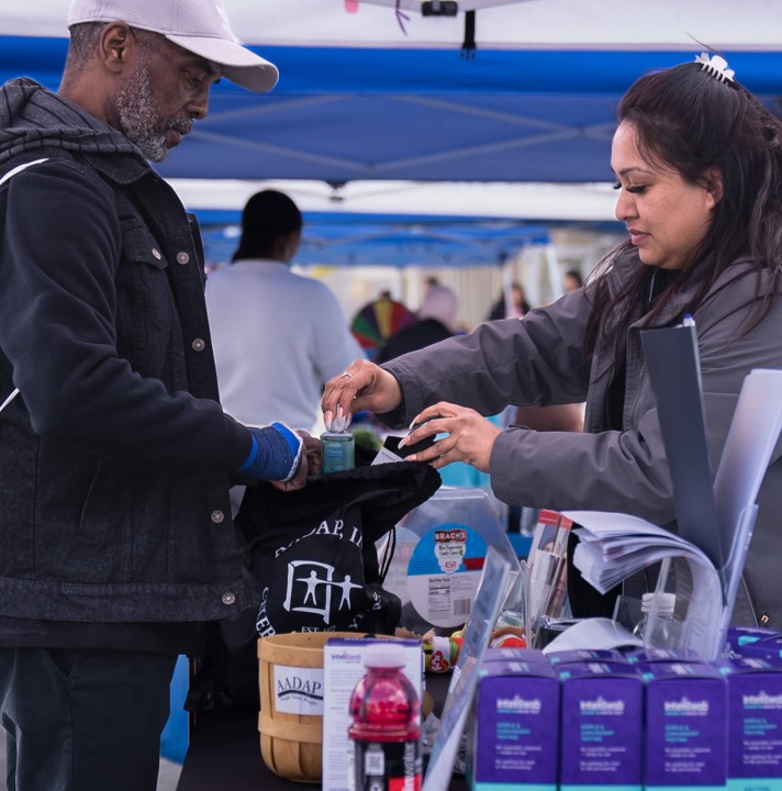 A woman places a bottle of hand sanitizer into a man's bag at a community event outdoors.