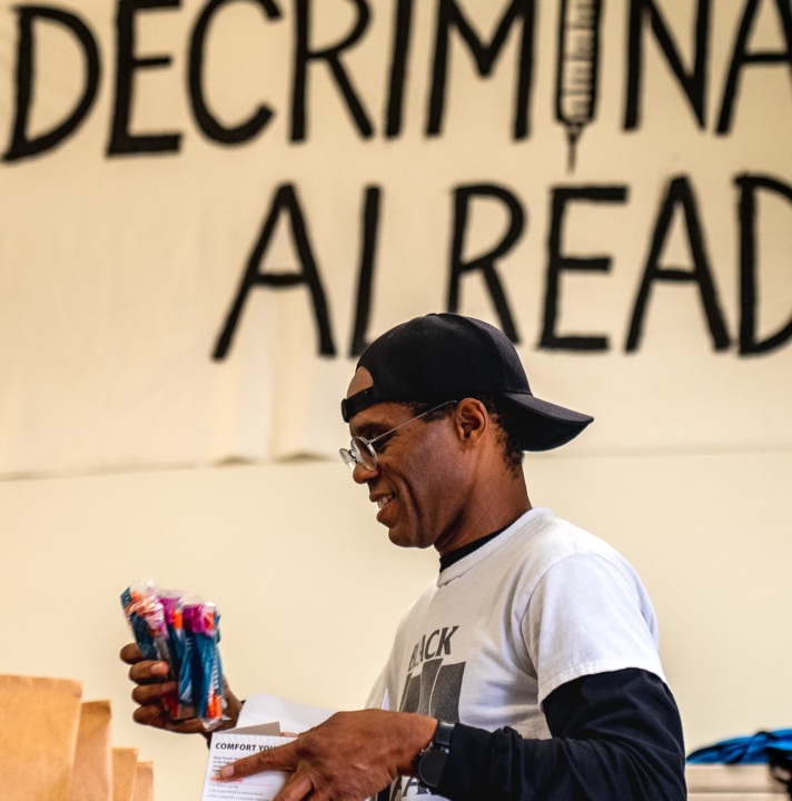 A Black man packs harm reduction kits. A banner behind him reads "Decriminalize Already".