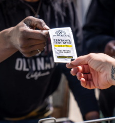 Hands exchange a fentanyl test strip packet. The packaging reads "Test before you ingest."