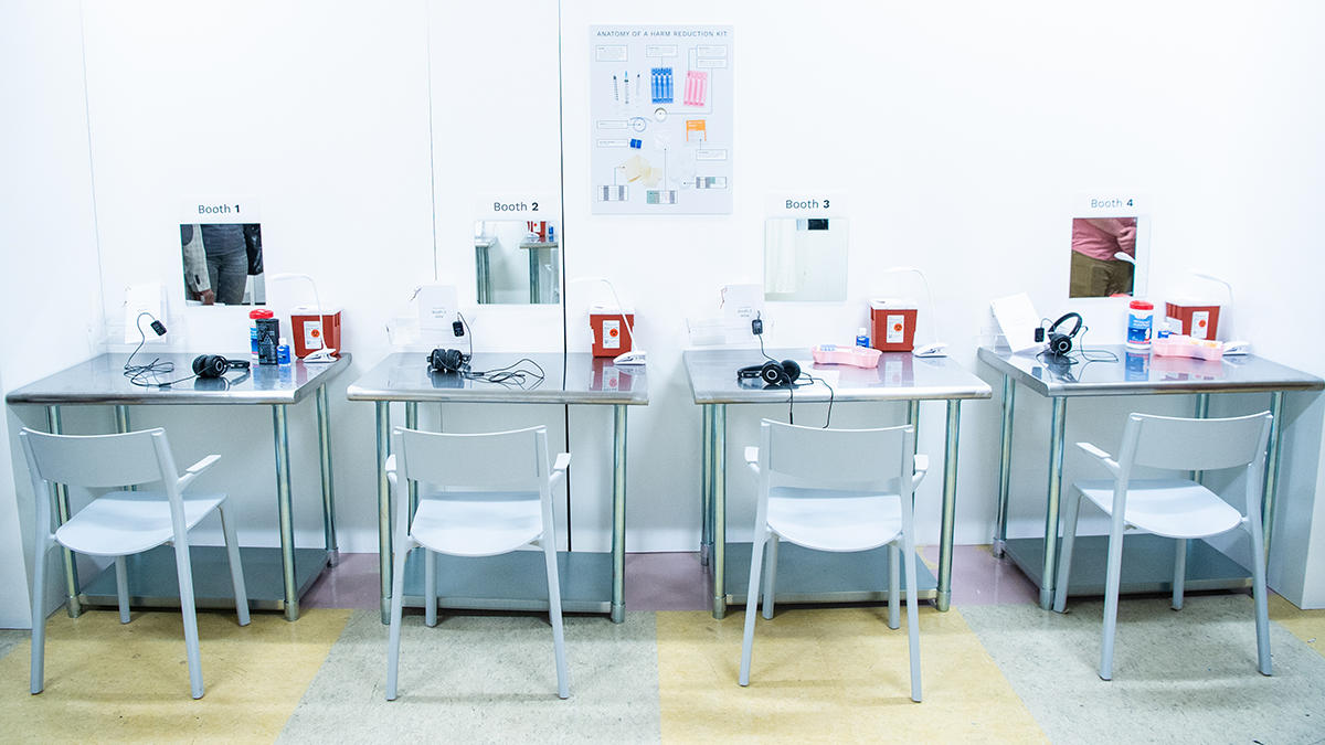 Booths with chairs and tables in a clean, bright overdose prevention center have harm reduction kits and supplies for visitors.
