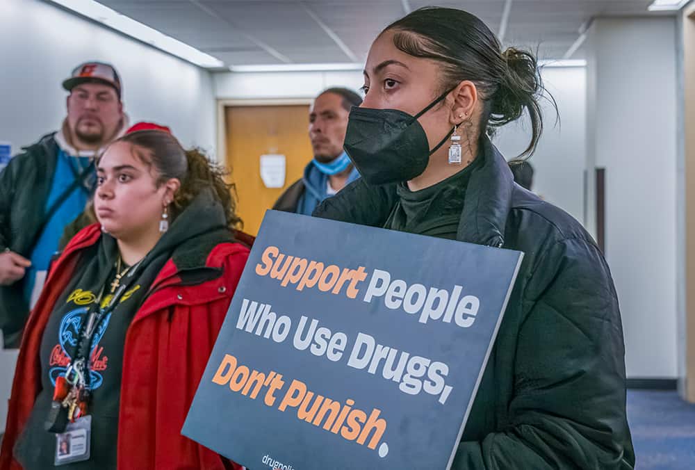 A woman holds a sign reading "Support People Who Use Drugs, Don't Punish."