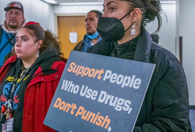 A woman holds a sign reading "Support People Who Use Drugs, Don't Punish."