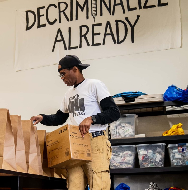 A man assembles harm reduction kits. A banner on the wall reads "decriminalize already".