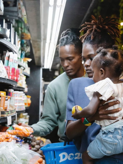 Photo of a black man, woman and child looking at a produce shelf. The man holds a bag of carrots. The woman is holding the child.