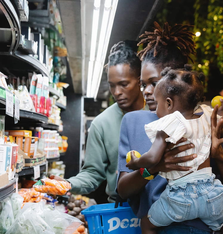 Photo of a black man, woman and child looking at a produce shelf. The man holds a bag of carrots. The woman is holding the child.