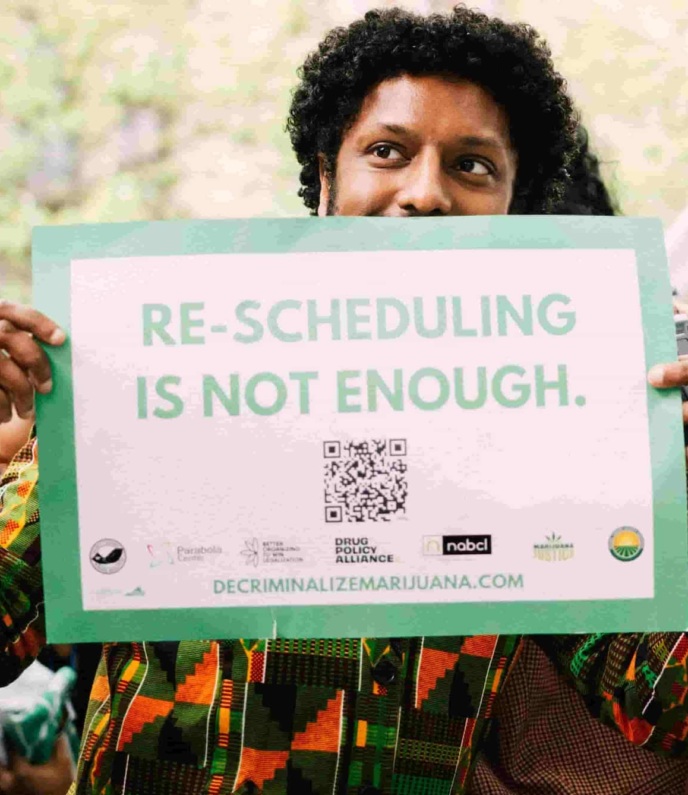 Photo of man holding a sign that says "Re-scheduling is not enough. decriminalizemarijuana.com"