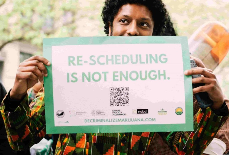 Photo of man holding a sign that says "Re-scheduling is not enough. decriminalizemarijuana.com"