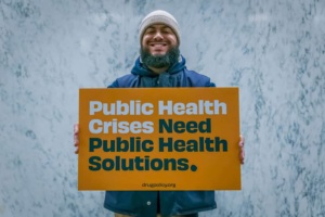 Photo of man holding sign that says "Public Health Crises Need Public Health Solutions. Drugpolicy.org"