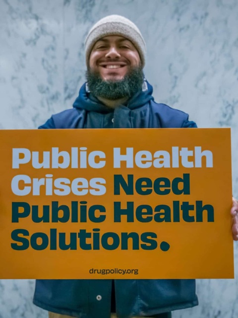 Photo of man holding sign that says "Public Health Crises Need Public Health Solutions. Drugpolicy.org"