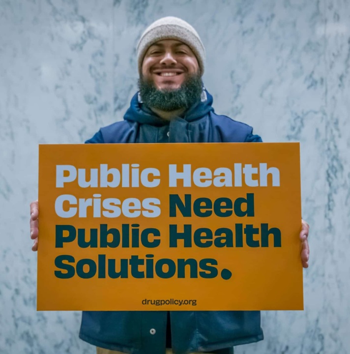 Photo of man holding sign that says "Public Health Crises Need Public Health Solutions. Drugpolicy.org"