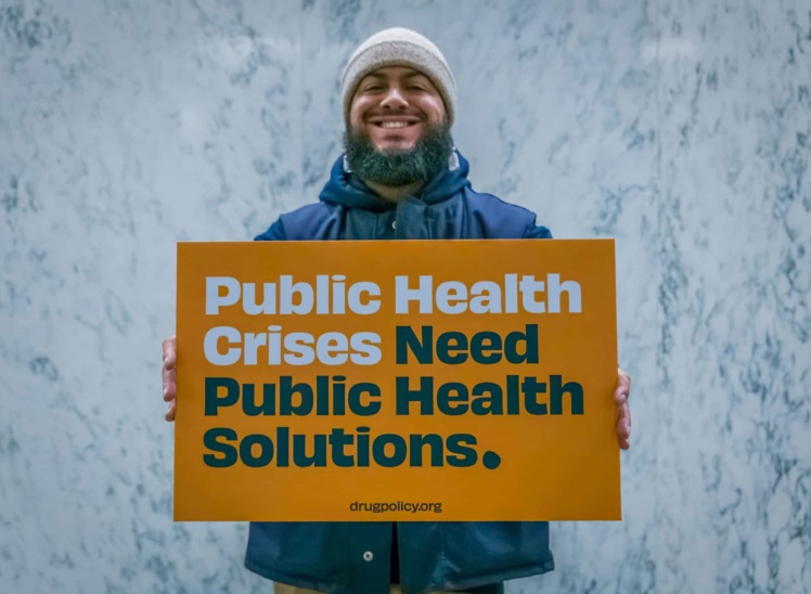 Photo of man holding sign that says "Public Health Crises Need Public Health Solutions. Drugpolicy.org"
