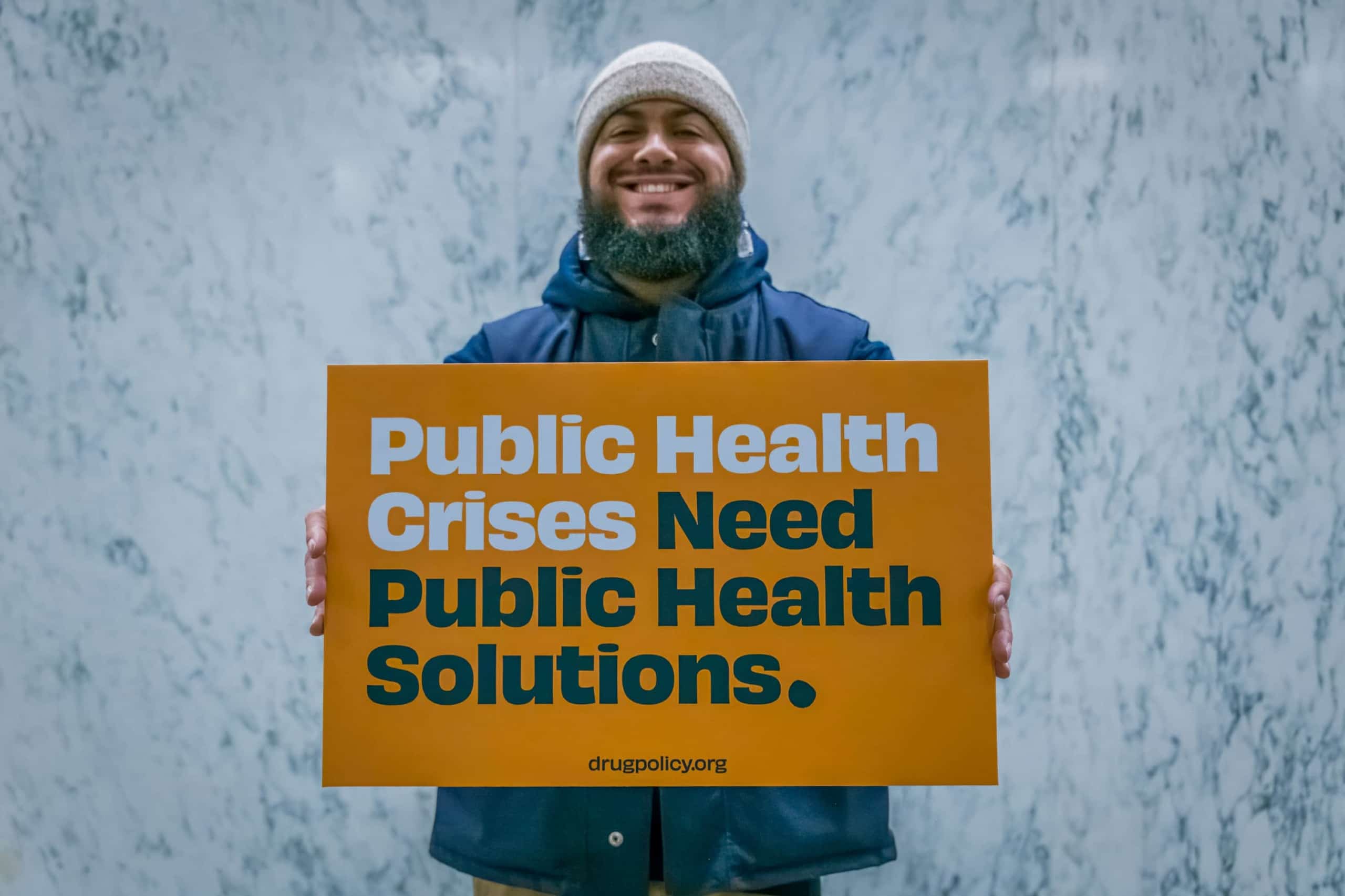 Photo of man holding sign that says "Public Health Crises Need Public Health Solutions. Drugpolicy.org"