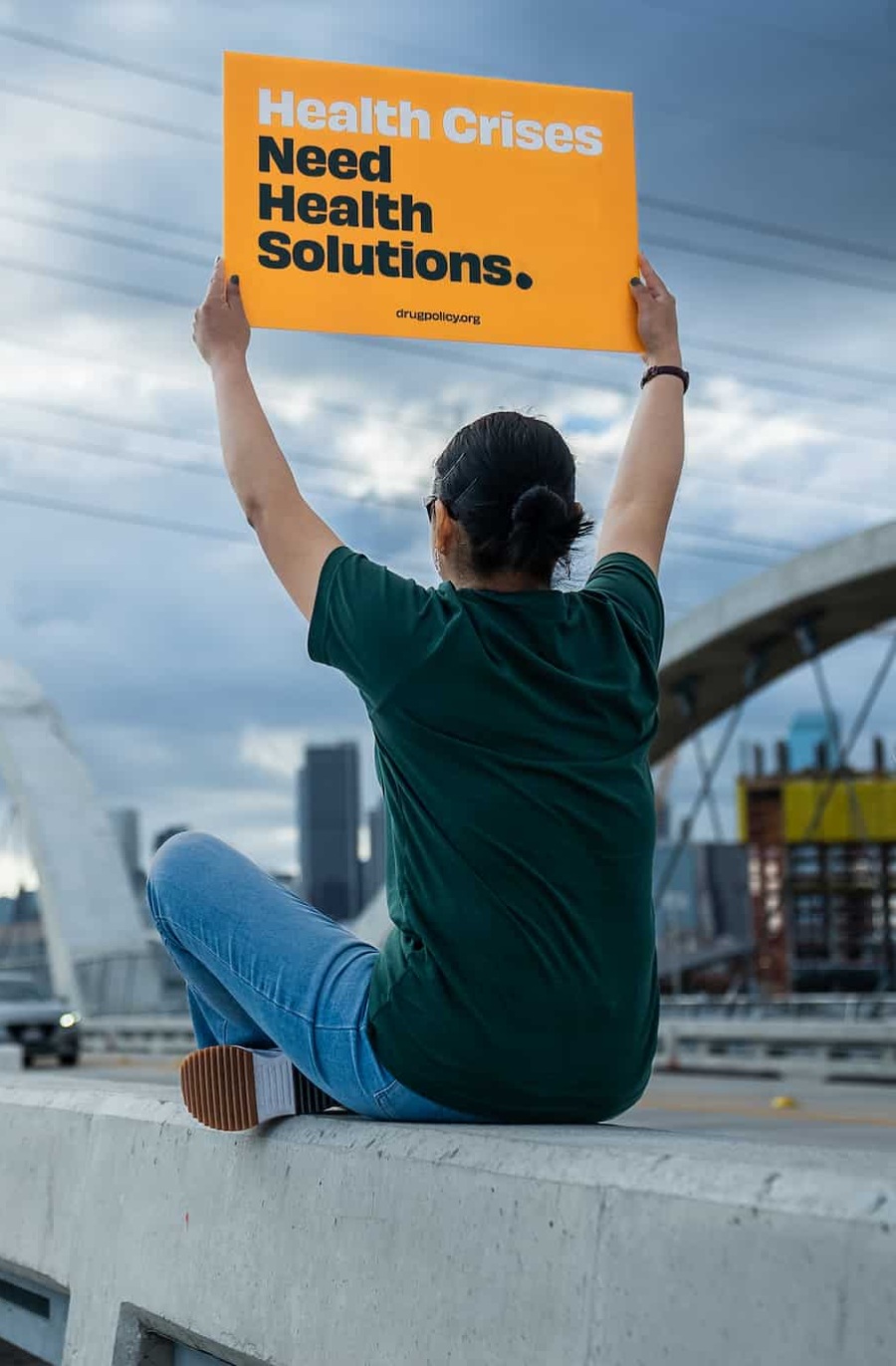 man sitting on a rooftop holding a sign saying "Health Crises Need Health Solutions"